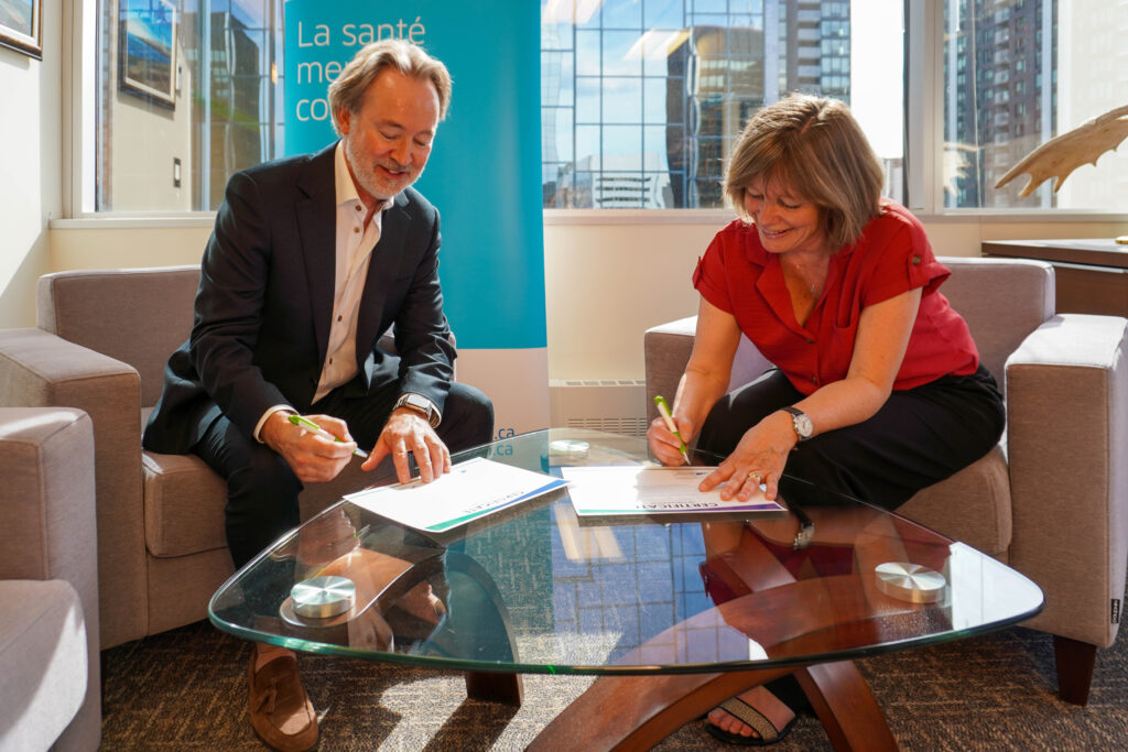 Seated in cushioned office lounge chairs, Michel Rodrigue, president and CEO at the Mental Health Commission of Canada, and AnnMarie Churchill, president and lead executive officer at Stepped Care Solutions, lean over a glass table to sign a certificate of partnership.
